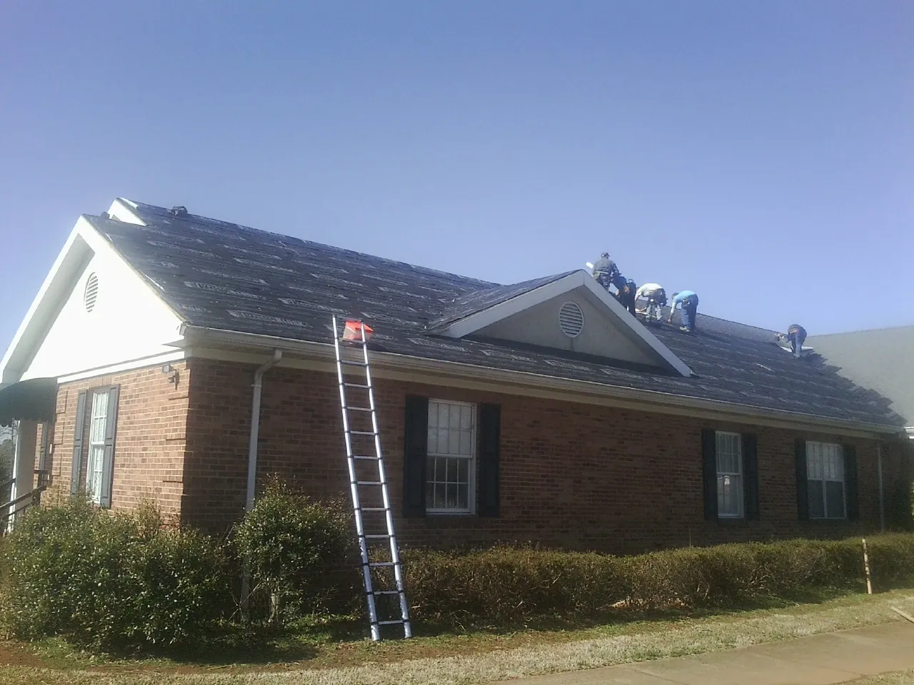 Crew tearing shingles off a brick ranch roof with a ladder set — Metal Roof Repair work by our crew in Orange Lake