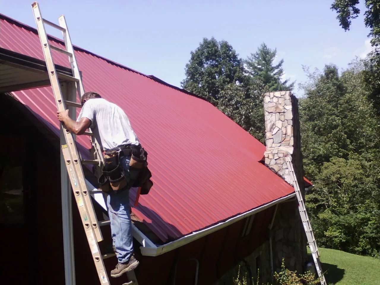 Roofer on a ladder beside a red metal roof and stone chimney — Soffit & Fascia Repair by Roofing Brothers in Orange Lake